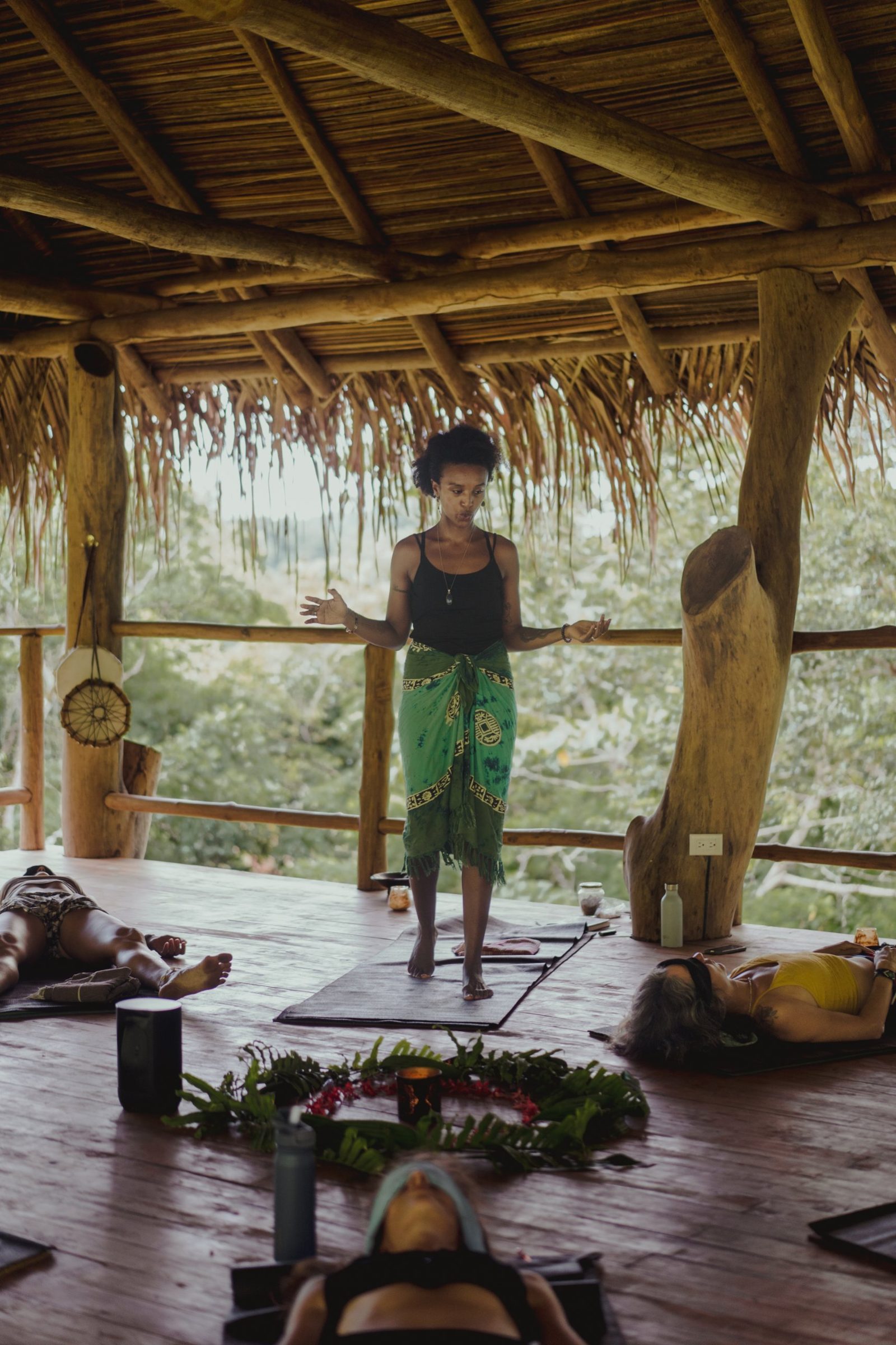 Badria guiding a group session under a thatched roof, participants resting on mats around a circle of leaves and flowers